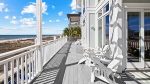 A wooden deck with white railings, a view of the beach, and a building with large windows. - Island Life: Beachfront home, Sleeps 28, Pool, Pet-friendly, All the amenities! (St. George Island)