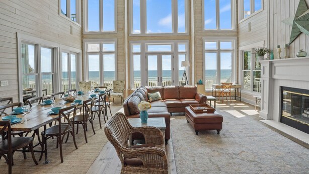 A spacious living area with a large window overlooking the sea, furnished with a sofa, chairs, and a dining table set for six. - Island Life: Beachfront home, Sleeps 28, Pool, Pet-friendly, All the amenities! (St. George Island)
