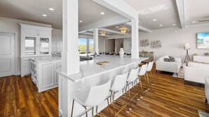 Eine moderne Küche mit weißen Schränken, einer großen Kochinsel mit Barhocker und einem Essbereich mit weißem Sofa und Stehlampe.