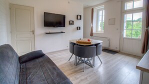 Una sala de estar con sofá, televisor montado en la pared, mesa de comedor con sillas y ventana con cortinas.