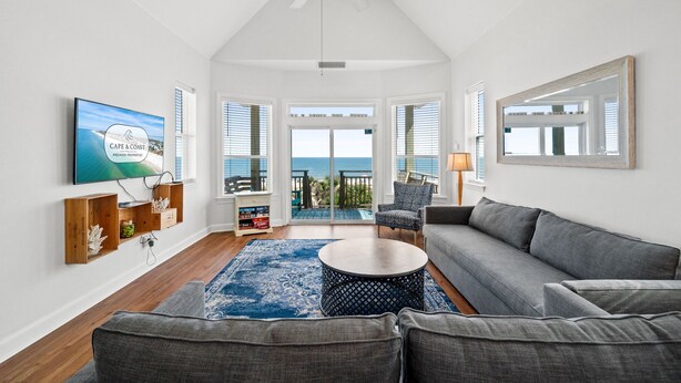 A spacious living room with a large gray sofa, a round coffee table, and a blue patterned rug. There is a television mounted on the wall, a ceiling fan, and a large window with a view of the ocean. - Above the Dune: 3 Bedroom, Beachfront, Sleeps 8 (St. George Island)