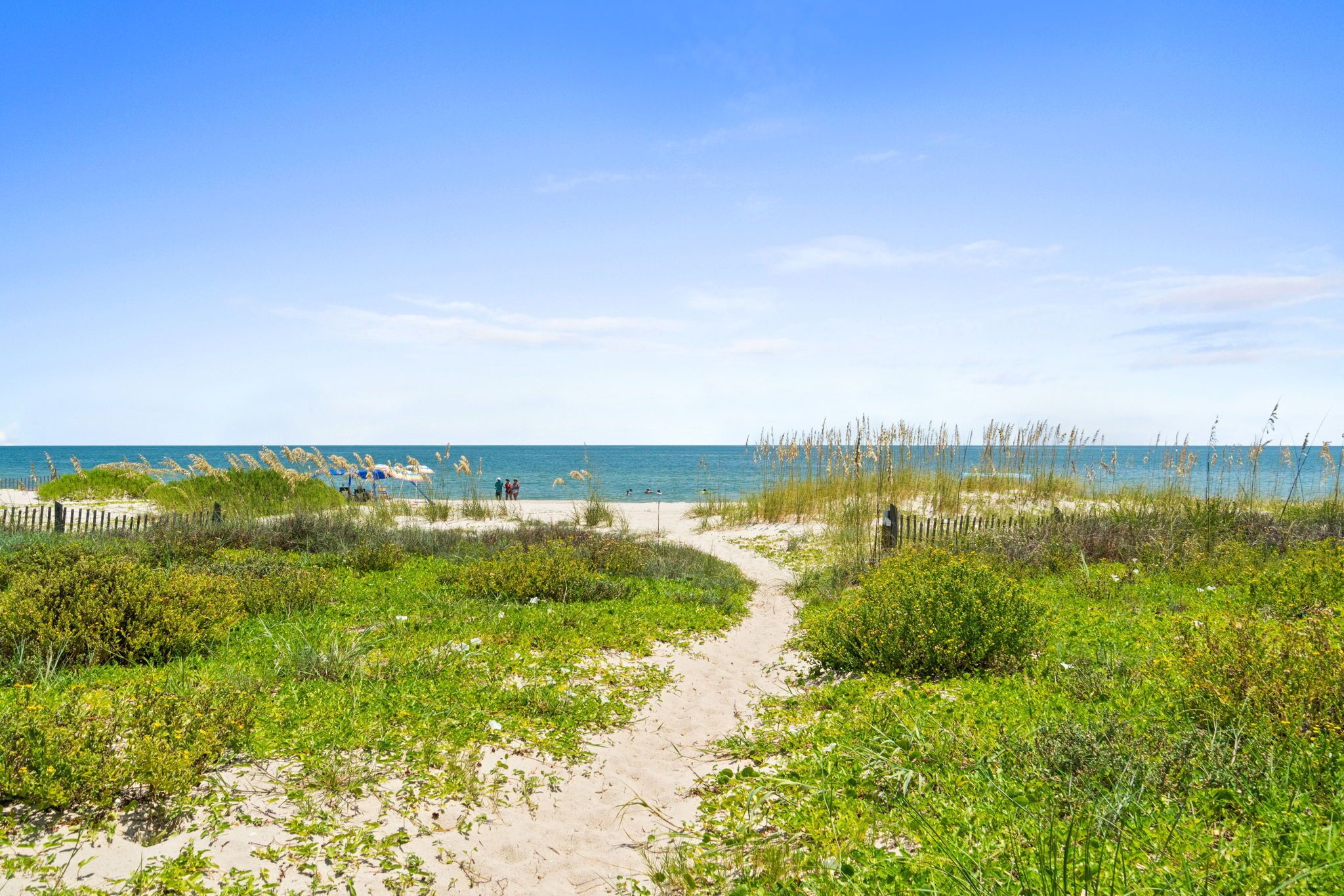 Un sentier de sable qui longe la plage et mène à l’océan, sous un ciel dégagé.