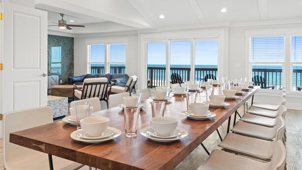 Ein langer Esstisch ist für eine Mahlzeit gedeckt, mit weißen Tellern und Gläsern, einem Holztisch und Blick auf das Meer durch große Fenster.