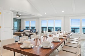 A long dining table set for a meal with white plates and glasses, a wooden table, and a view of the ocean through large windows. - Beach Blast: Luxury Beachfront Home, Private Pool, Pet-Friendly, Hot Tub (St. George Island)