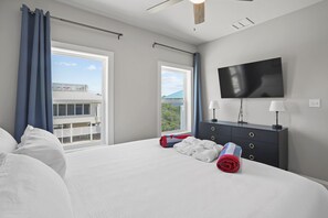 A hotel room with a large bed, two windows with curtains, a flat-screen TV, a dark dresser with two lamps, and a view of a building outside. - Blue Skies: Gulf Views, Private Pool, Pet Friendly (St. George Island)