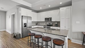 A modern kitchen with white cabinets, a granite countertop, stainless steel appliances, and wooden bar stools. - Blue Skies: Gulf Views, Private Pool, Pet Friendly (St. George Island)