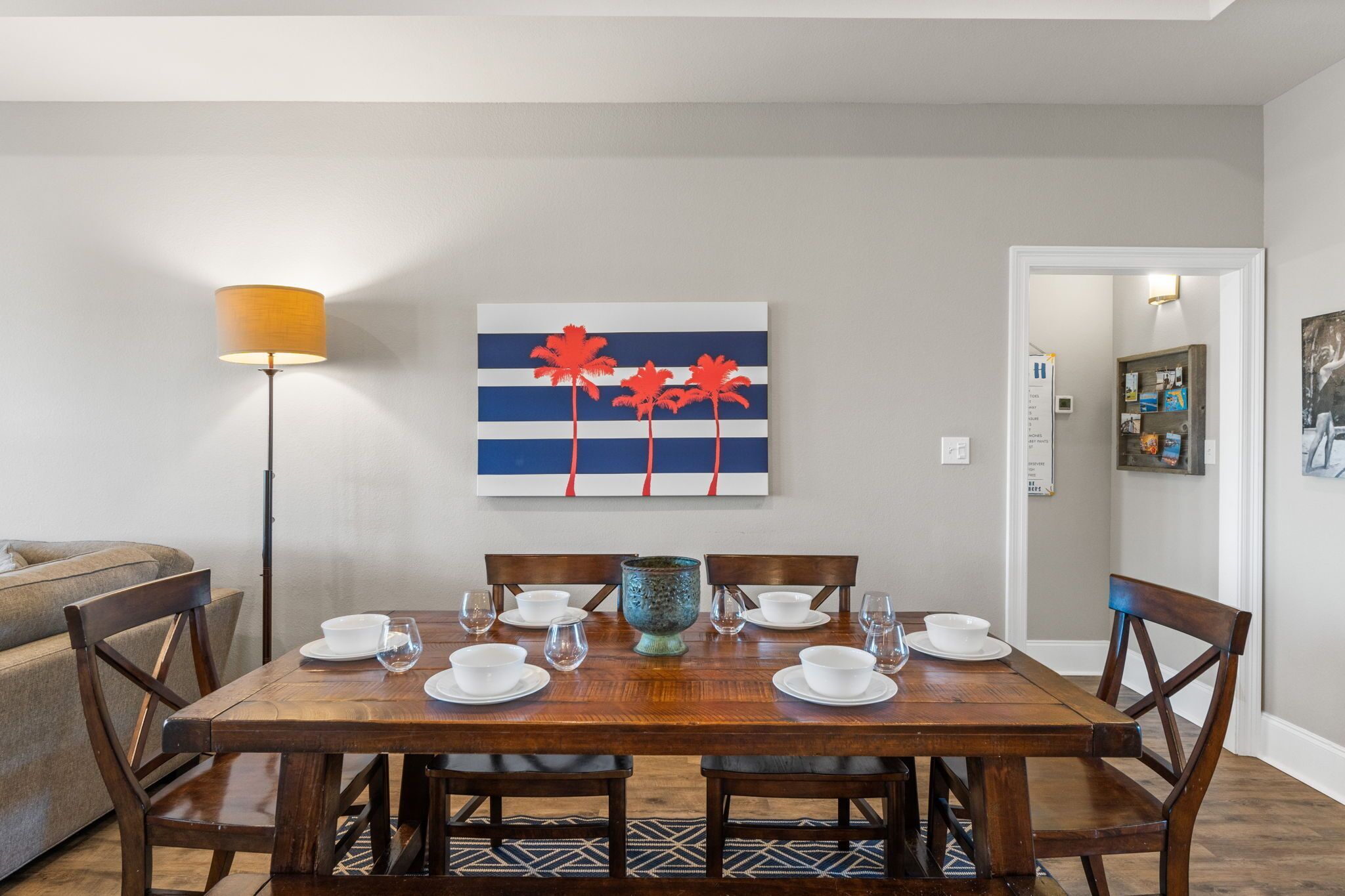 Een eethoek met een houten tafel voor vier personen, een vloerlamp en een schilderij van rode palmbomen aan de muur.