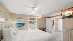Een slaapkamer met een bed, een plafondventilator, een aan de muur bevestigde televisie en een schilderij met een strandthema.