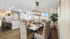 A dining area with a wooden table, wicker chairs, and a hanging light fixture. - Sugar Magnolia: Beachfront SGI Home- Sleeps 12, Priv. Pool, Pet Friendly! (St. George Island)