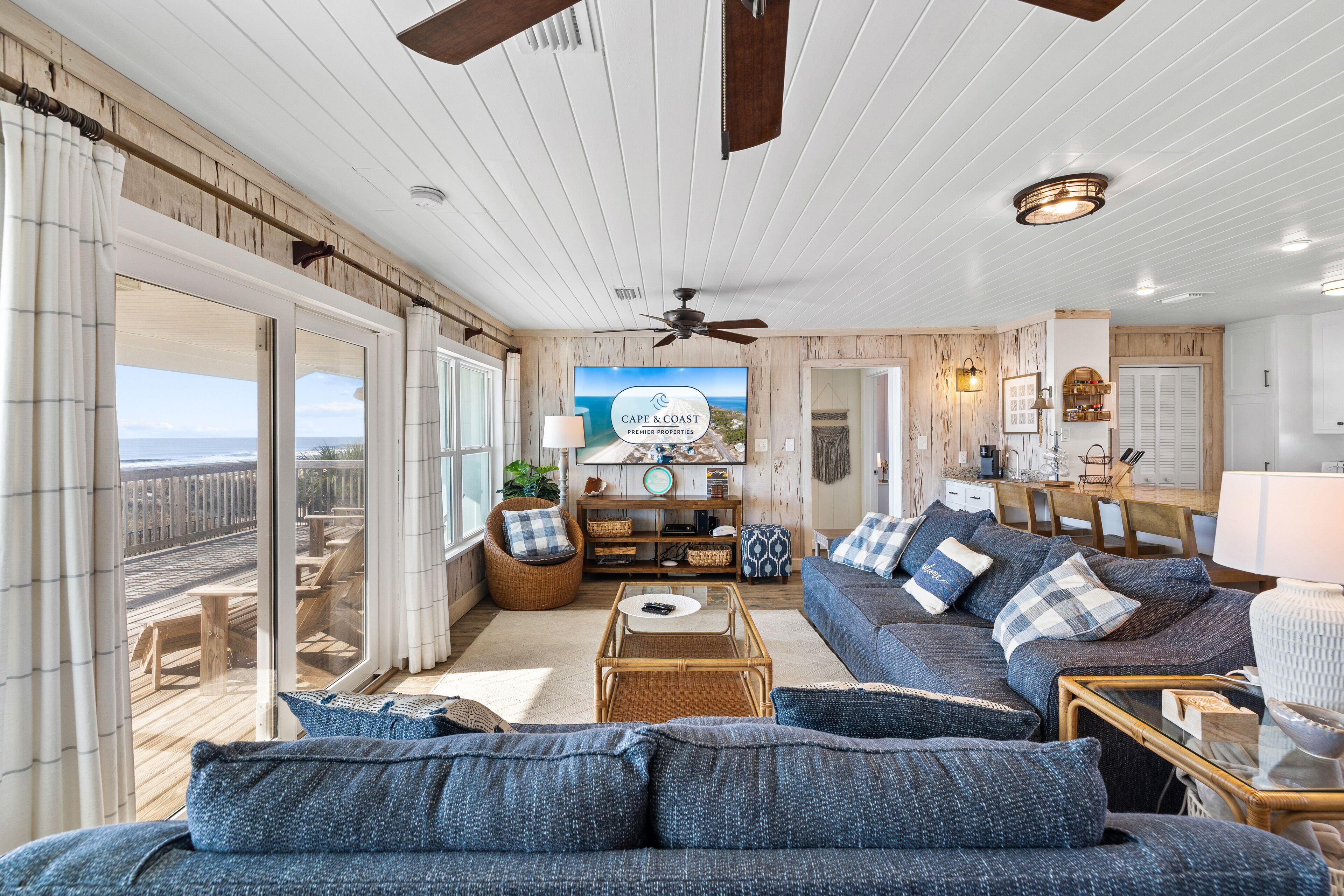 A spacious living room with a large television, comfortable seating, and a view of the ocean through large sliding glass doors.