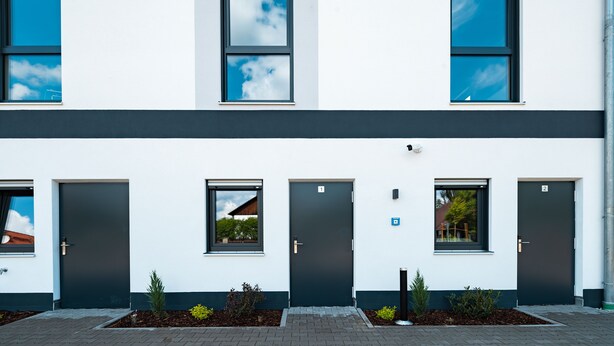 Un edificio moderno con fachada simétrica, destacando una puerta central flanqueada por ventanas y un pequeño jardín con plantas.
