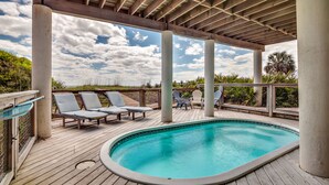 A wooden deck with a pool, lounge chairs, and a covered area with a view of the beach.