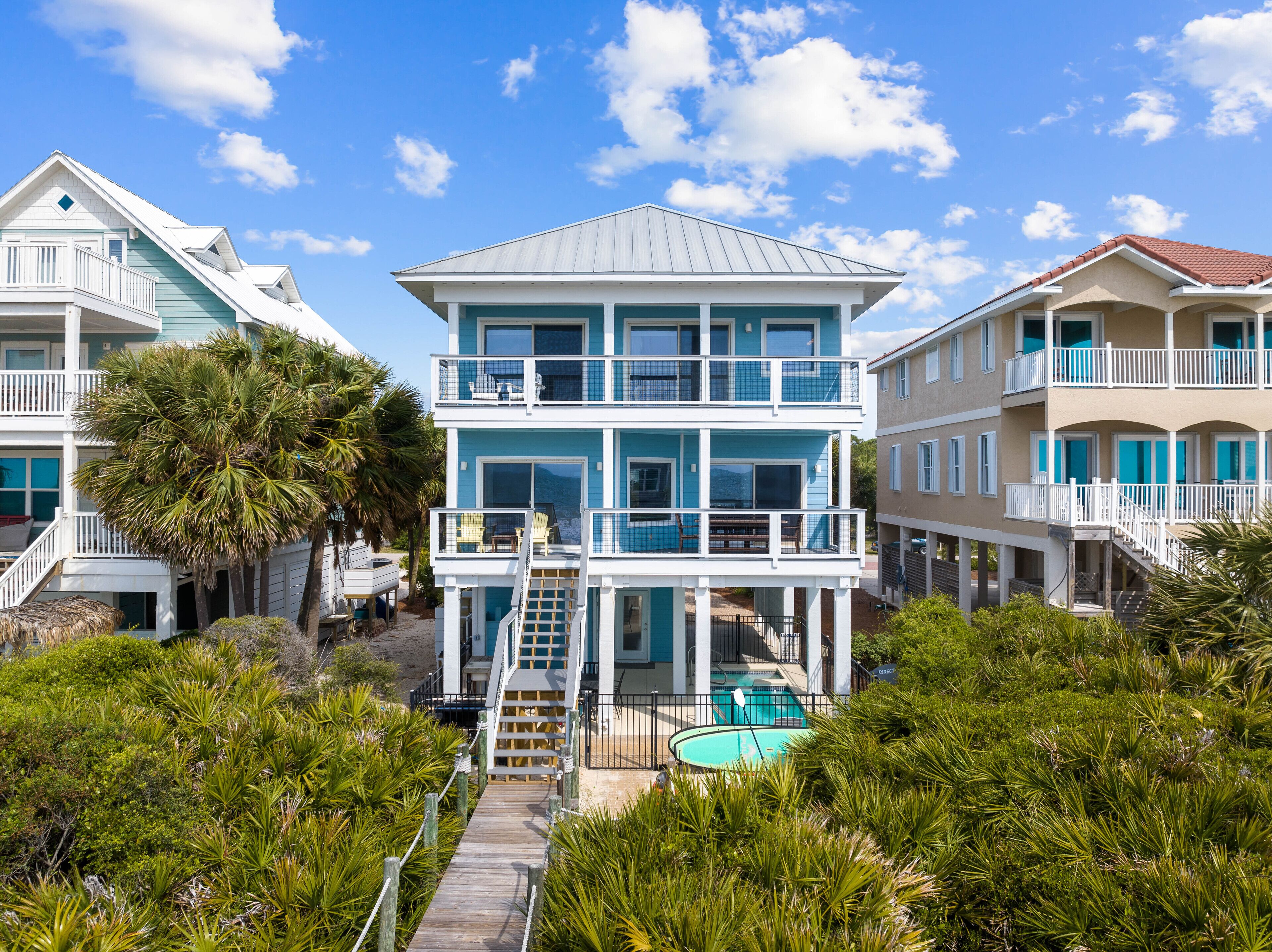 A multi-story beachfront house with a pool, balconies, and a wooden deck leading to the pool.