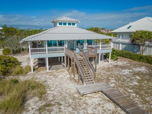 Une maison de plage dotée d’une terrasse couverte, d’un escalier menant à la terrasse et d’une plage de sable.