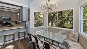 Una cocina moderna con estufa integrada, un comedor con mesa y sillas de vidrio, y amplias ventanas que ofrecen vistas a árboles.