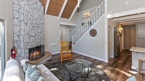 Amplia sala de estar con chimenea de piedra, una escalera de madera y una zona de cocina.