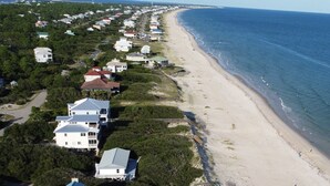 Ett flygbildsbild av ett bostadsområde vid kusten, med hus längs stranden, fri sikt över havet och en asfalterad väg som löper parallellt med stranden.