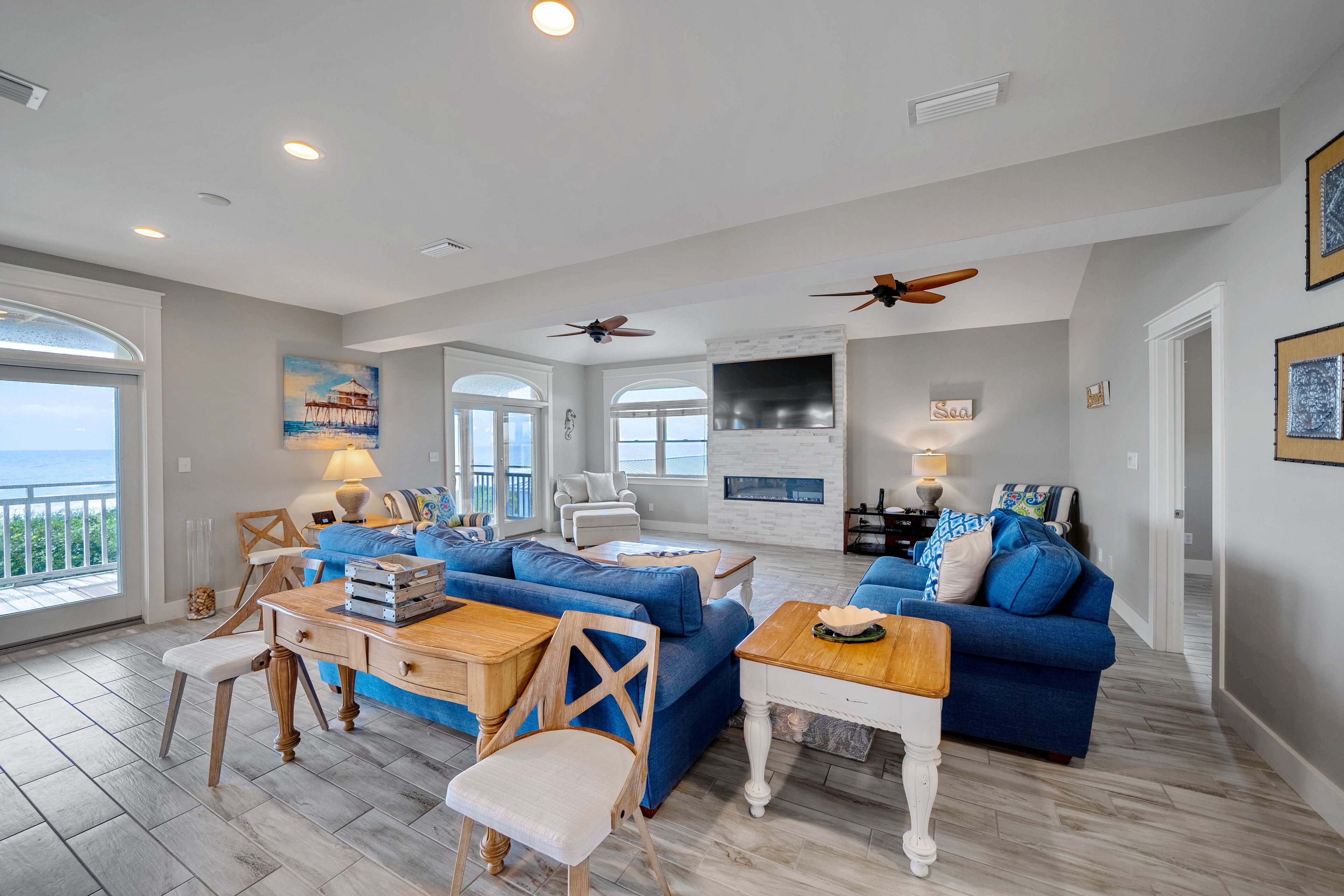 A spacious living room with a blue sofa set, wooden coffee table, and a dining table with chairs. There are ceiling fans, a fireplace, and a large TV mounted on the wall.