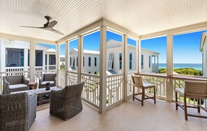 A covered outdoor area with wicker furniture, a table, and chairs, overlooking a coastal view.