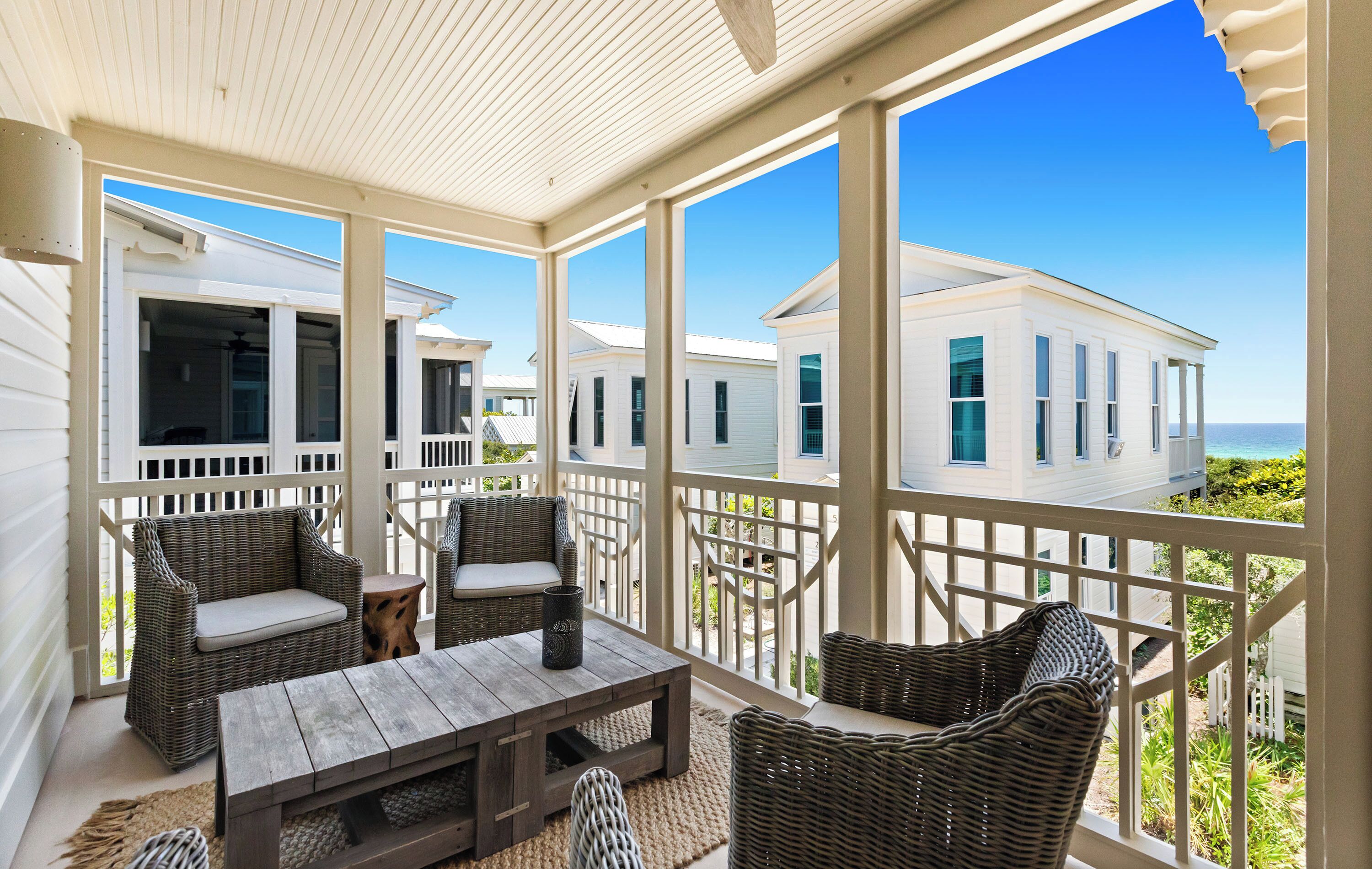 A covered outdoor area with wicker furniture, a wooden table, and a view of white buildings and the ocean.