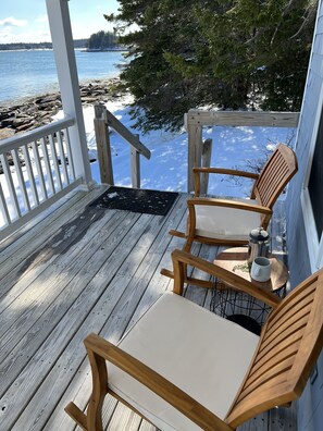 Eine Holzterrasse mit zwei Holzstühlen und einem Tisch, mit Blick auf einen verschneiten Strand und Wasser.