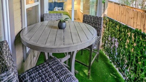 Katettu terassi, jossa on pyöreä pöytä ja punottuista materiaaleista valmistetut tuolit. Terassin ympärillä on puinen aita ja runsaasti viherkasveja.