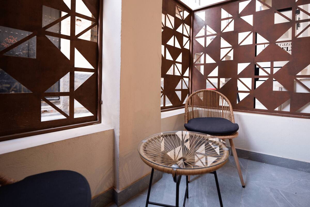 Une pièce avec un fauteuil en osier et une petite table ronde, un paravent en bois orné de motifs géométriques découpés, et une fenêtre donnant sur la rue.
