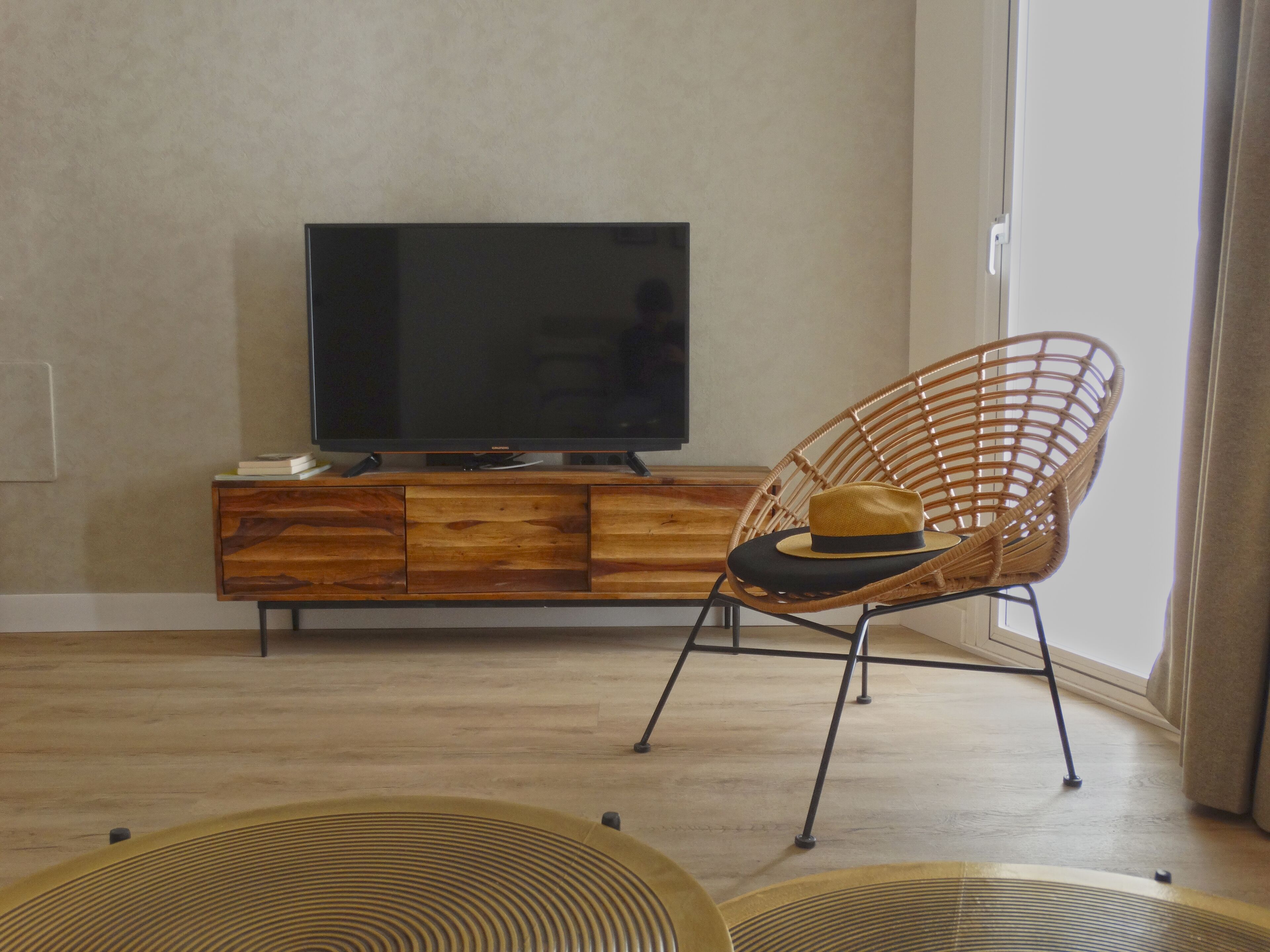 A living room with a wooden entertainment unit, a television, a wicker chair, and a large window.