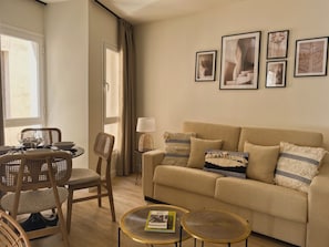 A living room with a beige sofa, two round tables, a dining table with chairs, and a window with curtains.