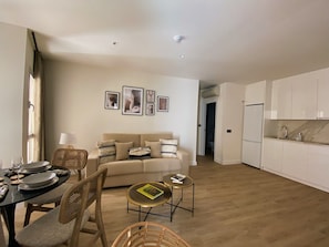 A modern living room with a sofa, dining table, and chairs, a kitchen with white cabinets, and a hallway leading to another room.