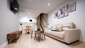 Un salon moderne avec un canapé, une table basse et une table d’appoint en bois. Un escalier mène à un espace cuisine doté de meubles blancs et d’une table à manger avec des chaises.