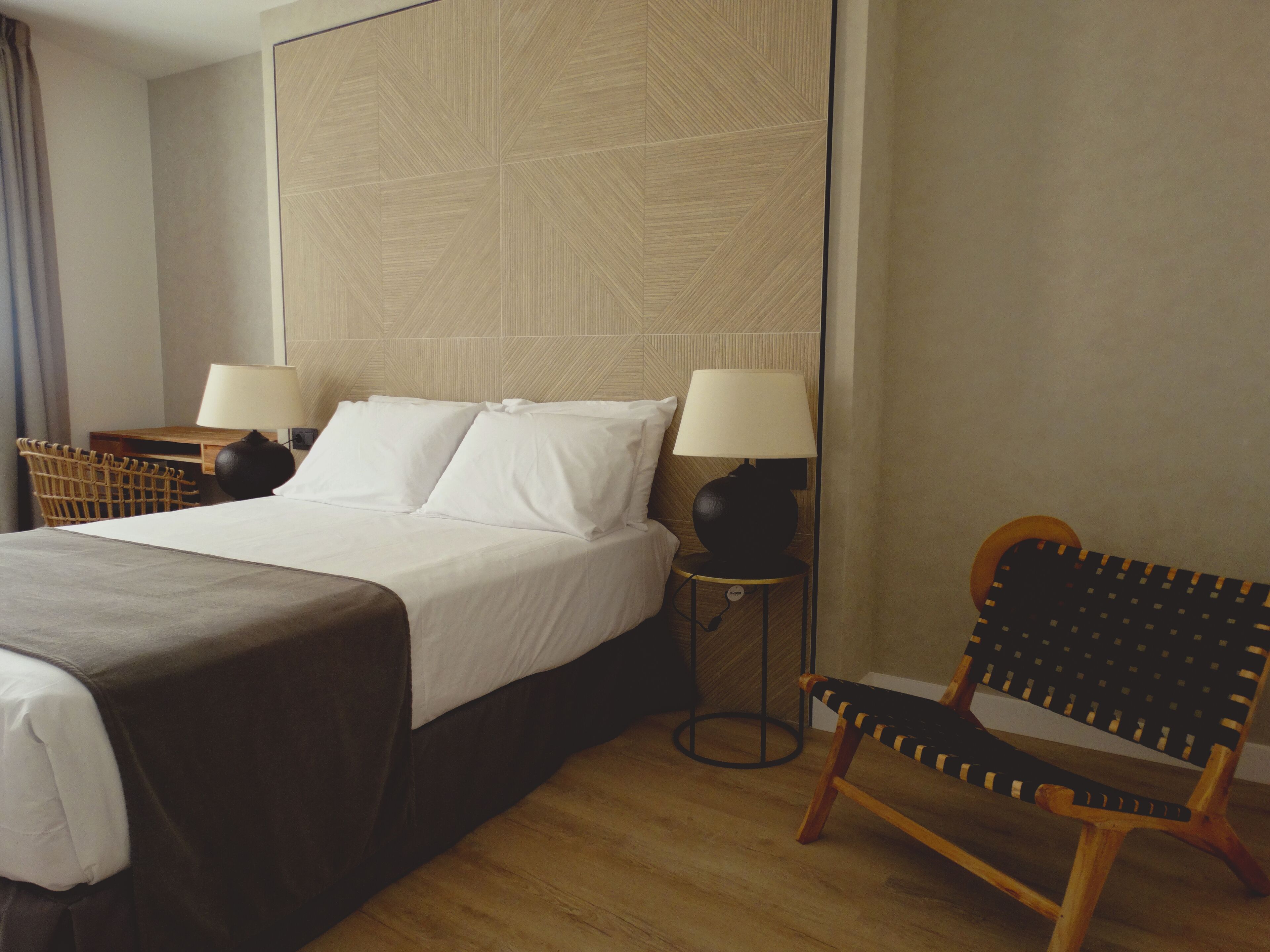 A neatly made bed with white linens, two bedside tables with lamps, a wooden chair, and a woven basket.