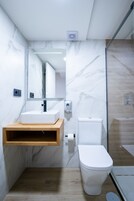 A modern bathroom with a marble wall, a wooden vanity, a white sink, a toilet, and a glass shower enclosure.
