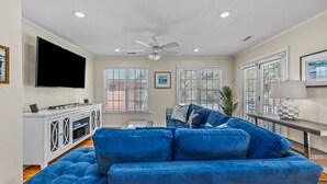 A living room with a blue sofa, a flat-screen TV, a ceiling fan, and a window with blinds. - Beach house w/ FREE Golf Cart & Fire Pit! (Pawleys Island)
