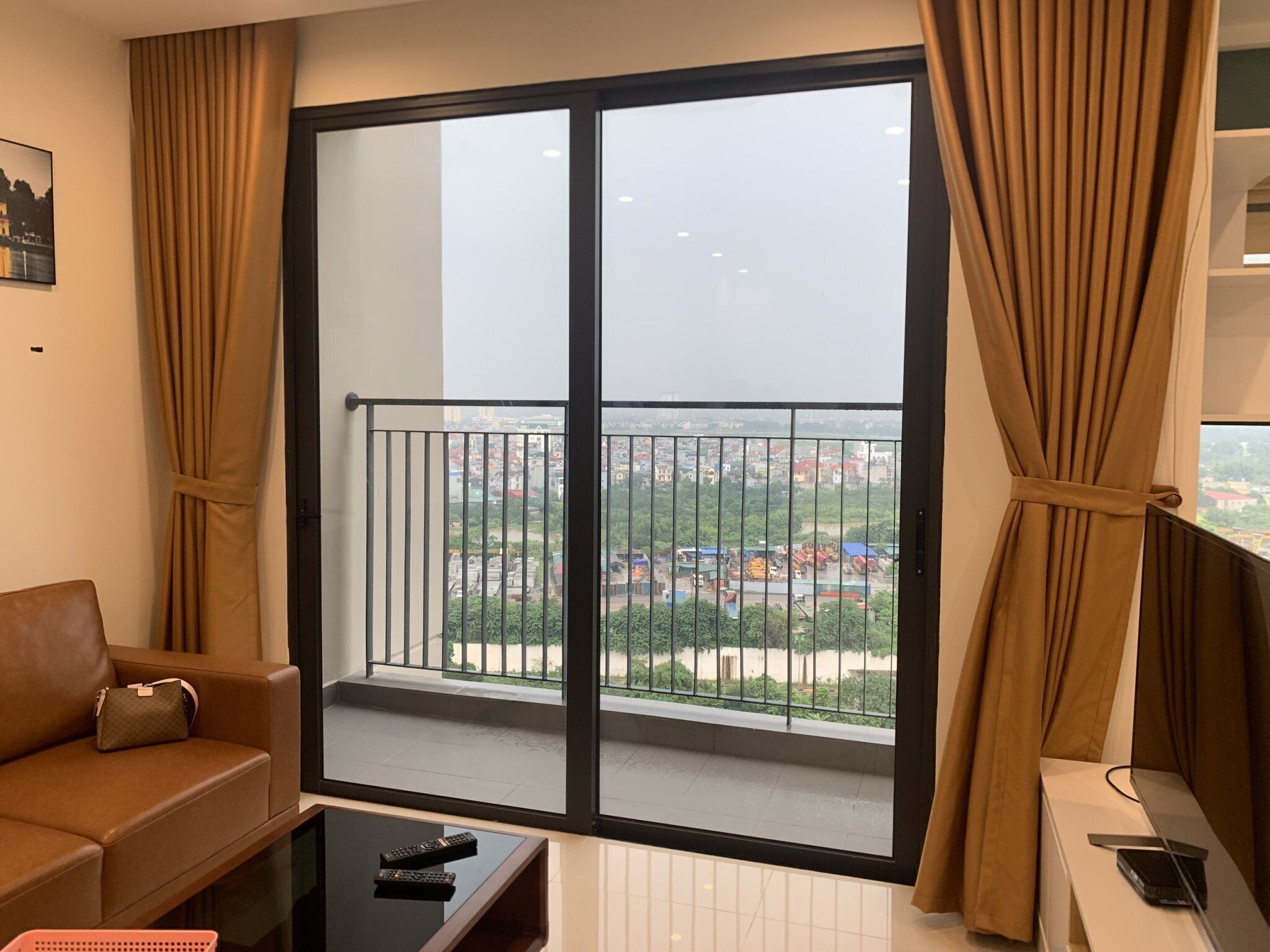 A modern living room with a large sliding glass door leading to a balcony, a brown leather sofa, a glass coffee table, and a TV on a stand.
