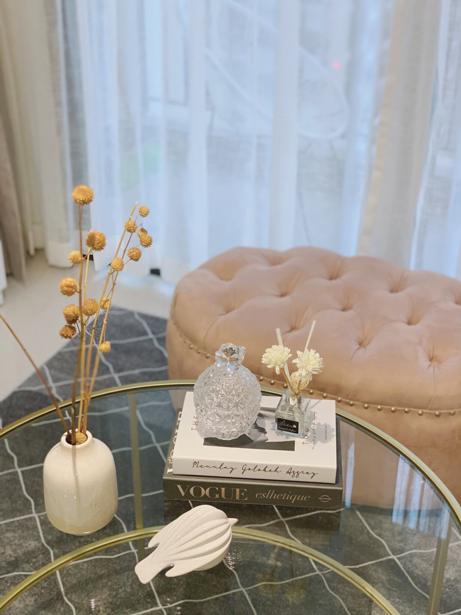A glass coffee table with a vase of dried flowers, a decorative glass bottle, and a book titled 'Vogue'.