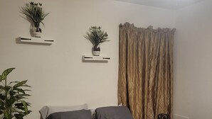 Cama bem arrumada com edredom cinza, uma pequena mesa de cabeceira com uma planta e duas prateleiras fixadas na parede com plantas em vasos.