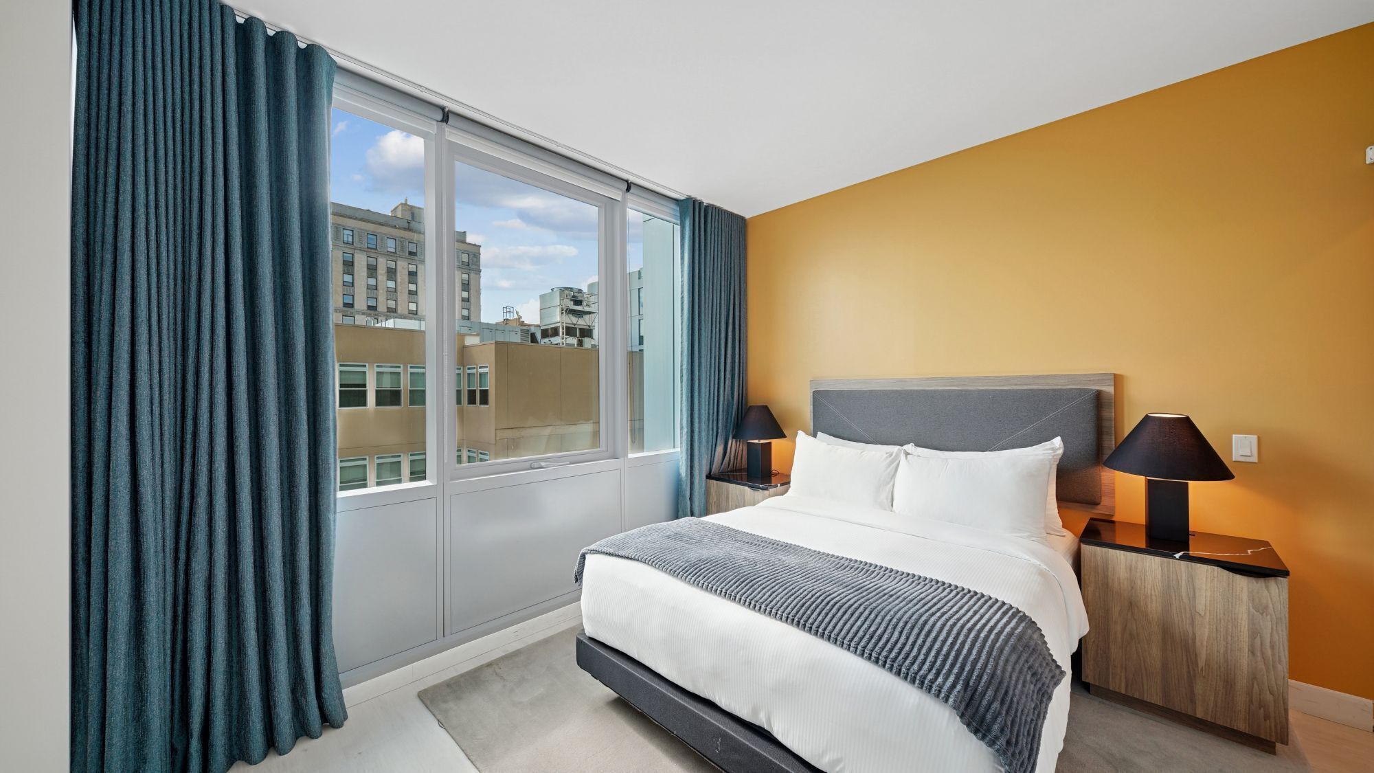 A hotel room with a large window, a bed with white bedding, two bedside lamps, and a view of buildings outside.