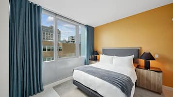 A hotel room with a large window, a bed with white bedding, two bedside lamps, and a view of buildings outside.