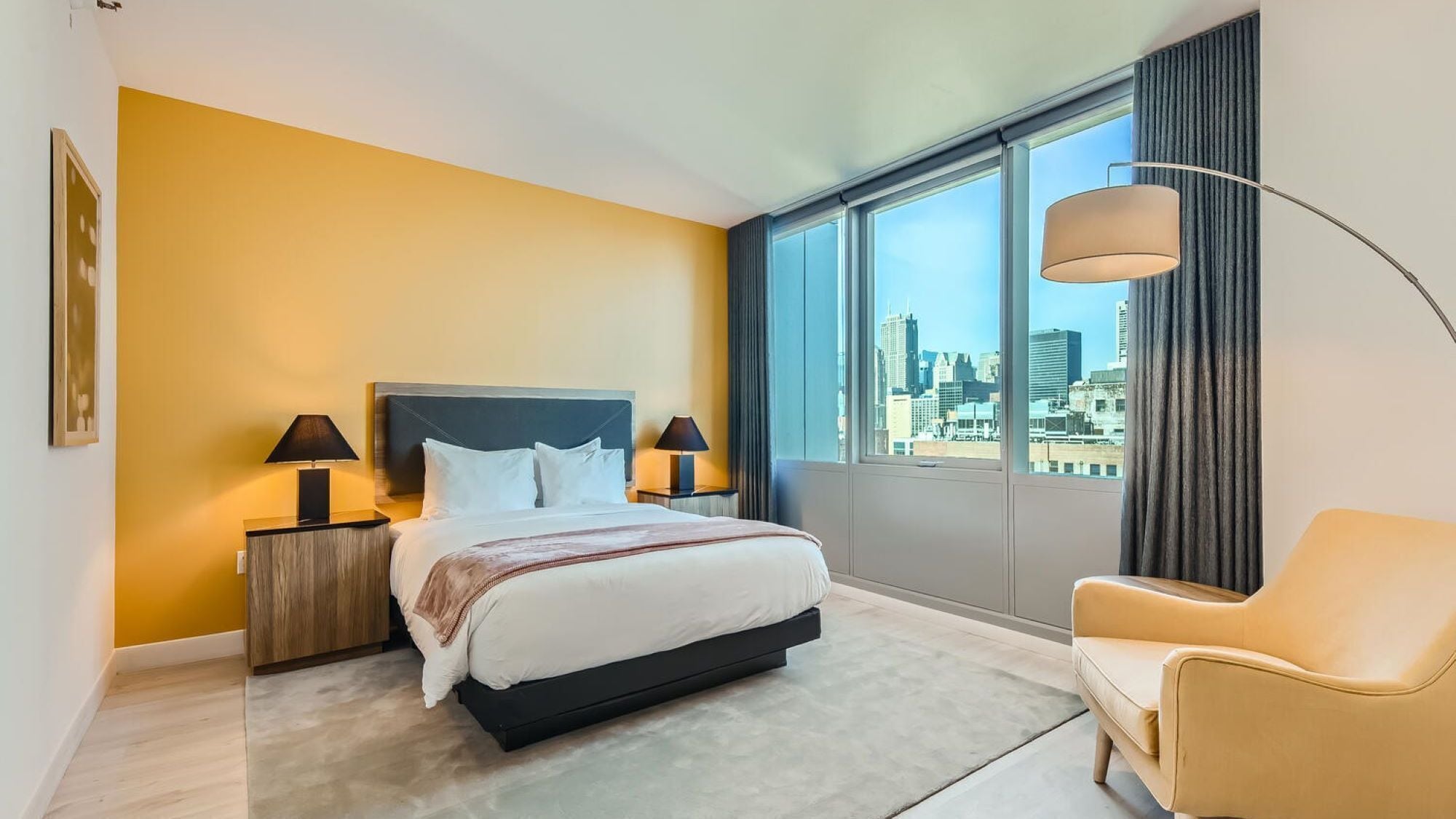 Ein Hotelzimmer mit einem Bett, zwei Nachttischen mit Lampen, einem Sessel, einem großen Fenster mit Stadtblick und einem Bild an der Wand.