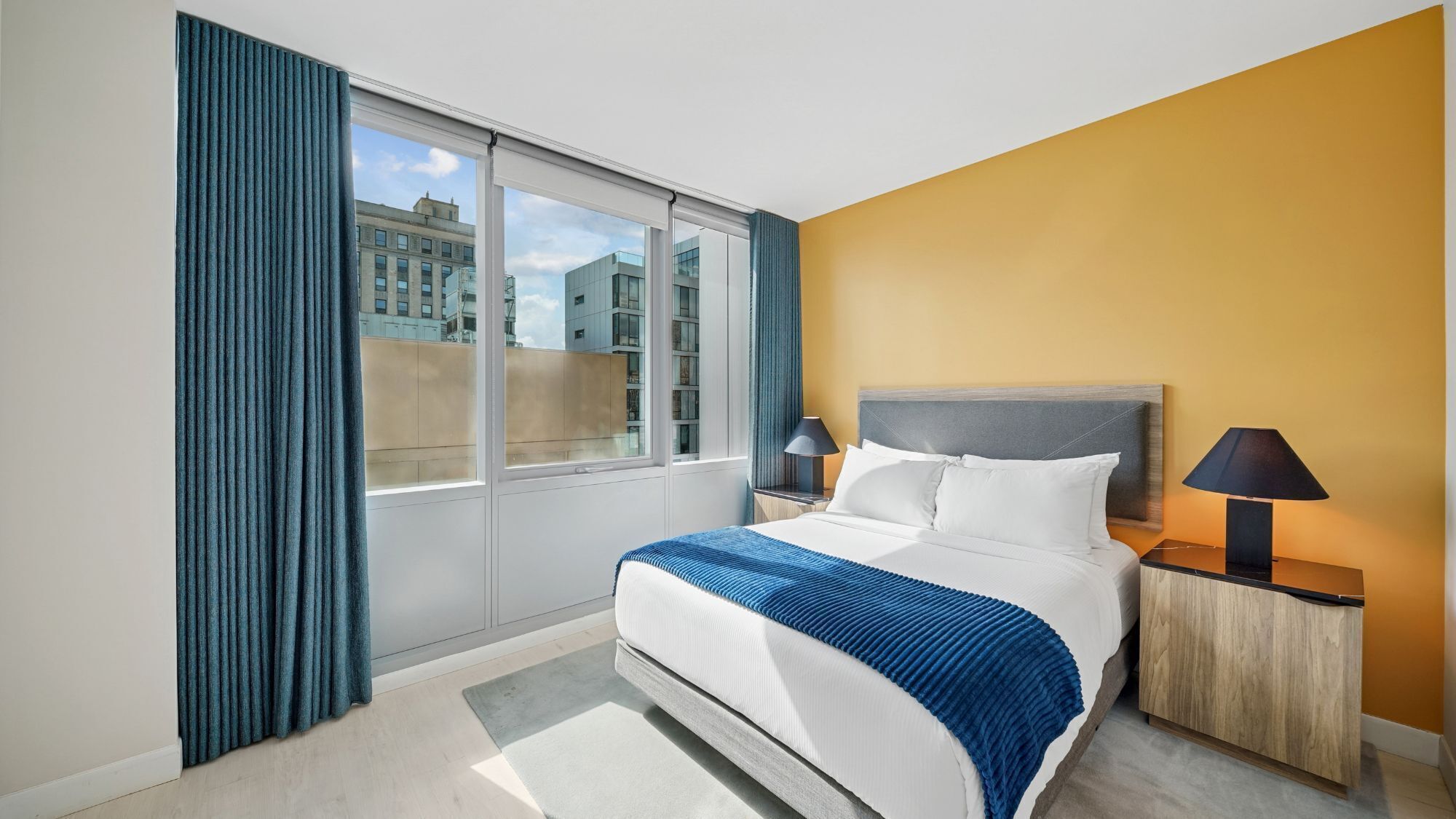 Una habitación de hotel con una cama, dos lámparas de noche, una ventana con cortinas y vistas a edificios.