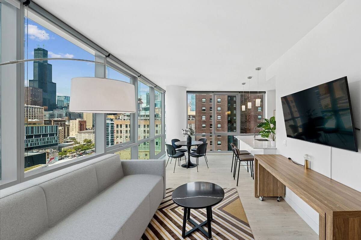 A modern living room with a large window, a sofa, a dining area, and a TV mounted on the wall.