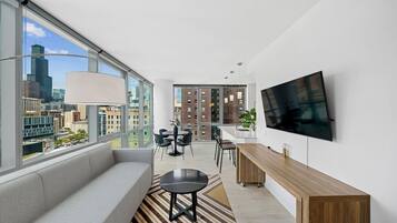 A modern living room with a large window, a sofa, a dining area, and a TV mounted on the wall.