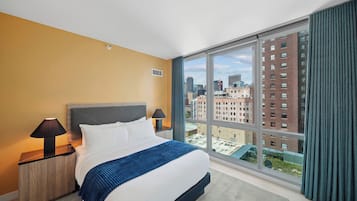 Une chambre d’hôtel avec un grand lit, une table de chevet avec une lampe, une vue sur la ville à travers de grandes fenêtres et un panorama urbain visible à l’extérieur.