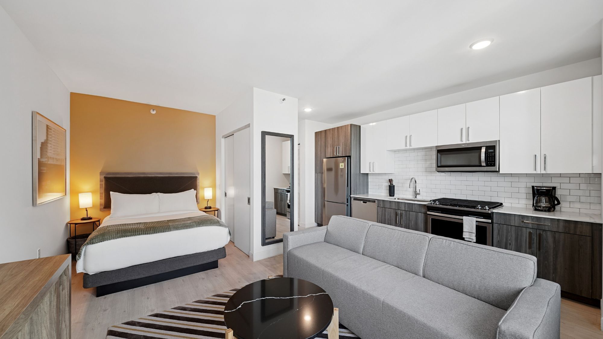 Una habitación de hotel moderna con una cama, un sofá gris, una mesita negra pequeña, una cocina con armarios blancos, un microondas y un refrigerador.