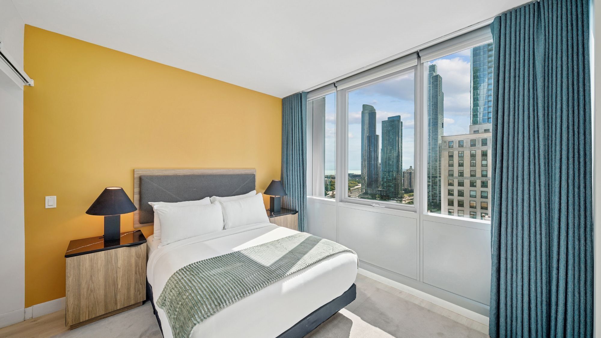 Habitación de hotel con cama, mesitas de noche, lámpara, vista de edificios altos y una ventana con cortinas.