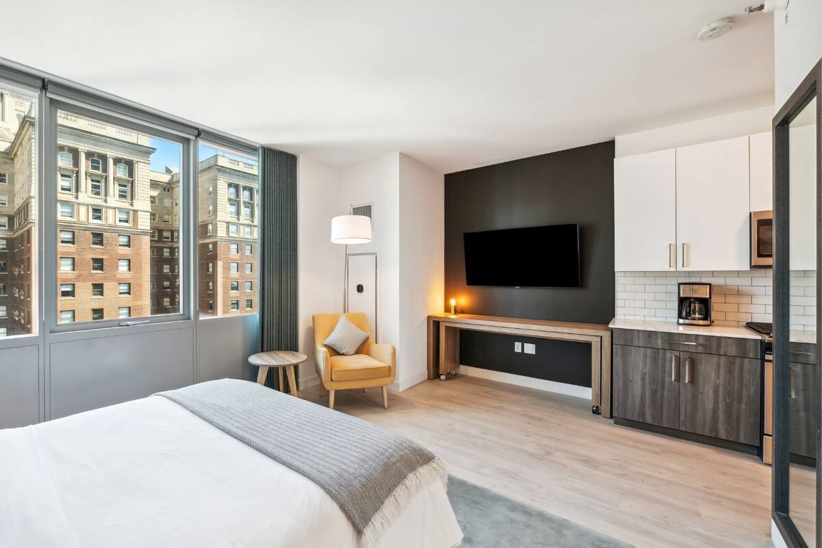 A hotel room with a large window, a bed, a chair, a TV, and a kitchenette.