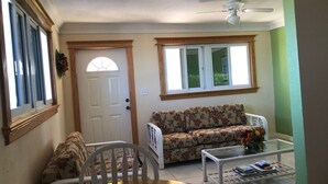 A living room with a floral sofa, a dining table with chairs, a coffee table with books, a ceiling fan, and a door with a window.