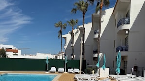 Zona junto a la piscina con sillones, palmeras y un cielo azul despejado.