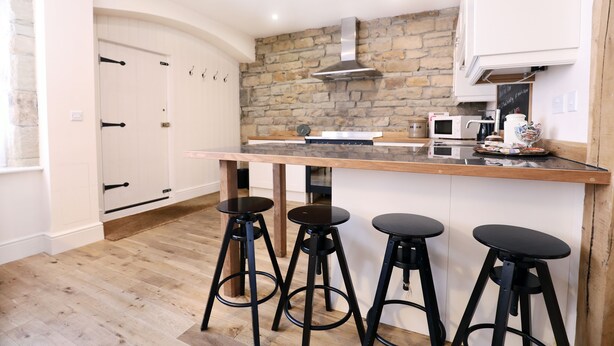 A modern kitchen with a stone wall, wooden countertop, and three black stools. - Stable Cottage (Harrogate)